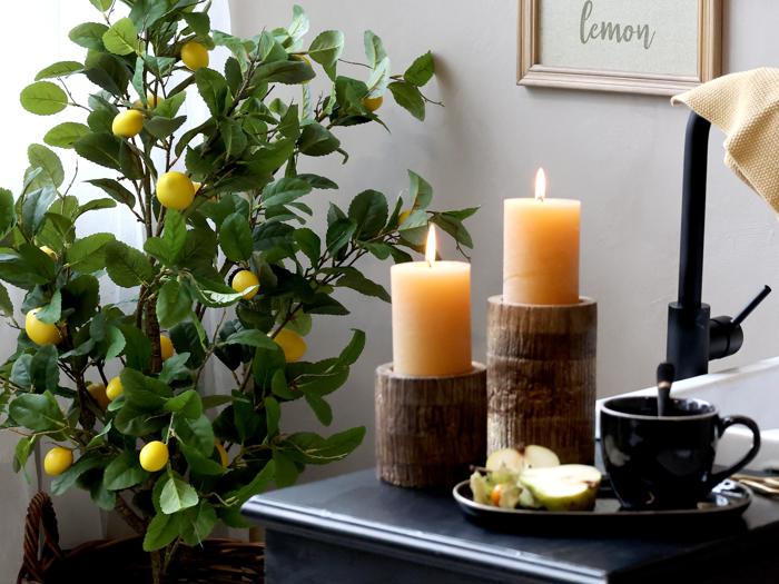 To tændte stearinlys på rustikke stager ved siden af en sort kop, underkop og frugtskiver med et citrontræ og et indrammet citronskilt i baggrunden.