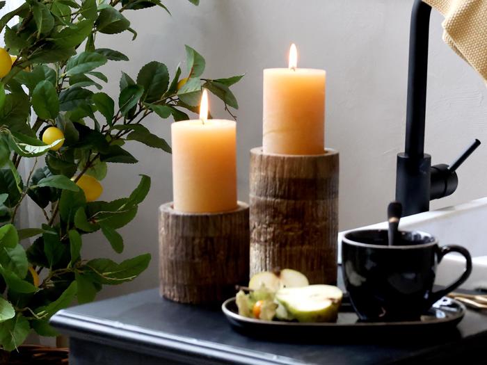 To tændte bloklys i træholdere står ved siden af en sort kop med en ske og en tallerken med udskåret frugt, i nærheden af en bladplante og en sort vandhane.