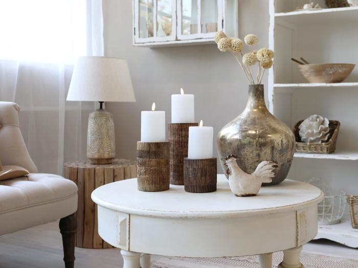 En hyggelig stue med et hvidt rundt bord med tre tændte bloklys, en metalvase med tørrede blomster og en keramisk kylling. Neutrale toner og naturligt lys fylder rummet.