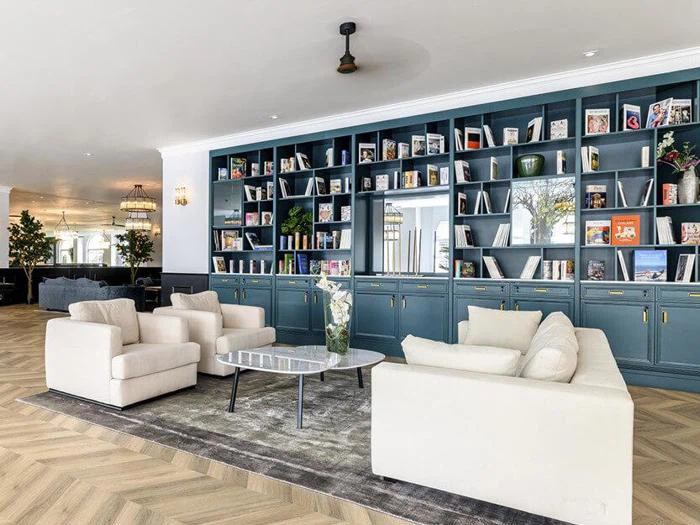 Moderne lounge med cremefarvede sofaer, et sofabord, en stor indbygget bogreol fyldt med bøger og indretning, lyst trægulv og lys, luftig belysning.