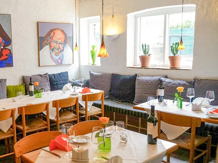 Hyggelig restaurant med træstole, dækkede borde, farverige servietter, vinflasker, blomster, pottekaktus i vindueskarmen og et portræt af en smilende mand på den hvide væg.