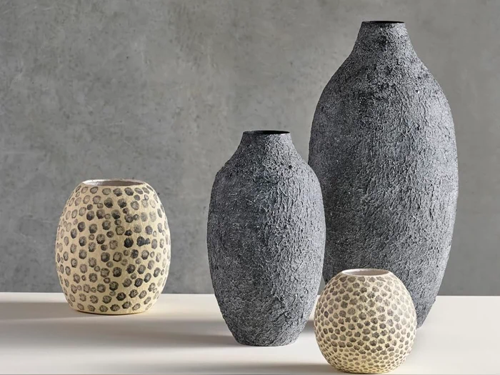 Fire teksturerede vaser i forskellige størrelser, to mørkegrå og to cremefarvede med prikkede mønstre, er udstillet på en lys overflade mod en grå baggrund.