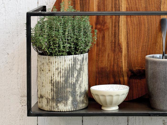 En timianplante i potte, en lille hvid skål og en grå kop står på en sort metalhylde foran et skærebræt af træ.