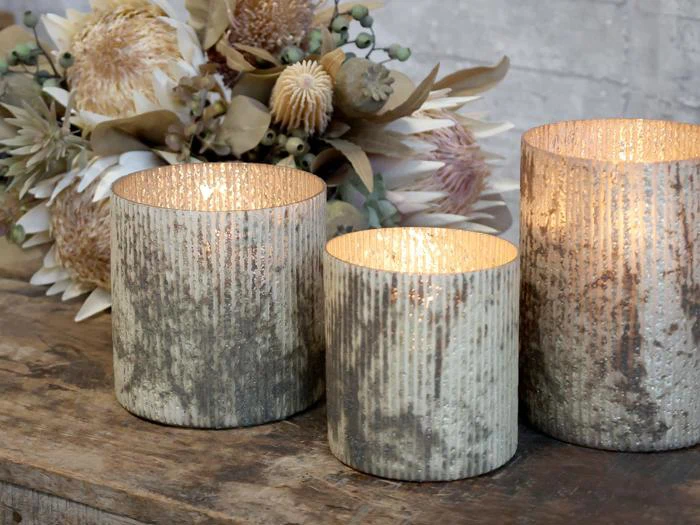 Tre teksturerede glaslys med varmt lys står på et rustikt træbord ved siden af en buket tørrede blomster og blade.