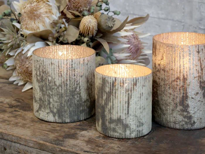 Tre teksturerede, metalliske lysestager med tændte stearinlys indeni står på en rustik træoverflade med tørrede blomster i baggrunden.