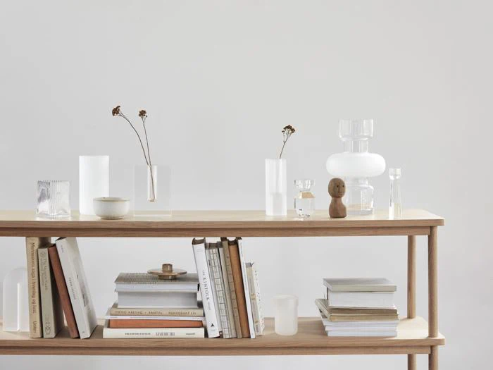 En minimalistisk træhylde rummer bøger, stablet og oprejst, sammen med klare glasvaser, en lille træfigur og et par tørrede blomster i et lyst, neutralt rum.
