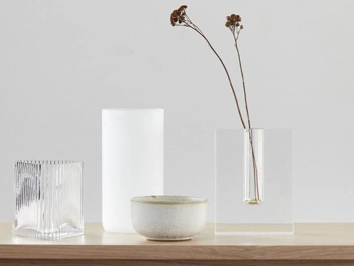 Fire minimalistiske beholdere - riflet glas, matteret glas, keramikskål og akrylvase med tørrede blomster - er arrangeret på en lys træoverflade mod en ensfarvet baggrund.