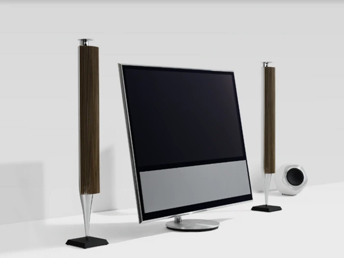 Et moderne fladskærms-tv med en sølvfarvet og sort bund er flankeret af to høje, slanke højttalere med en lille subwoofer i baggrunden på en hvid overflade.