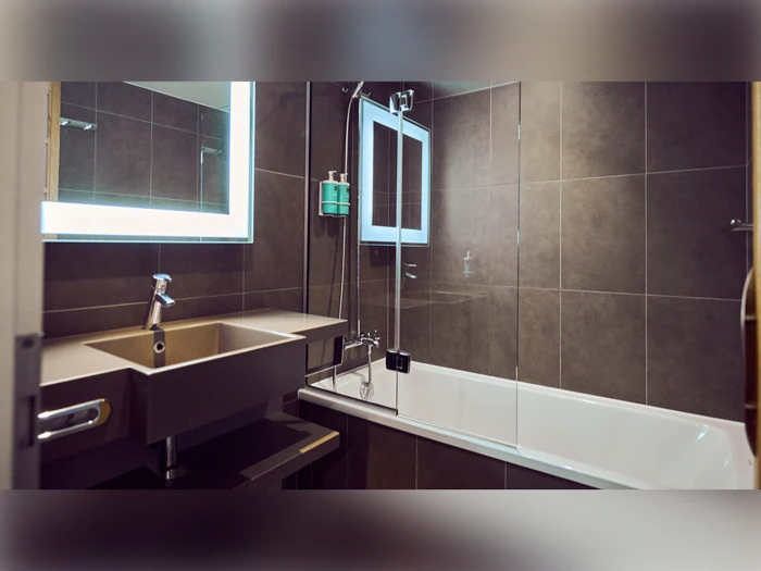 Moderne badeværelse med oplyst spejl, håndvask og badekar. Væggene er flisebelagte, og der er et monteret brusehoved med en glasskillevæg over karret.