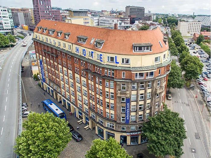 Et stort murstenshotel med rødt tag og blåt HOTEL-skilt ligger på et byhjørne omgivet af træer, veje, parkerede biler og bybygninger.