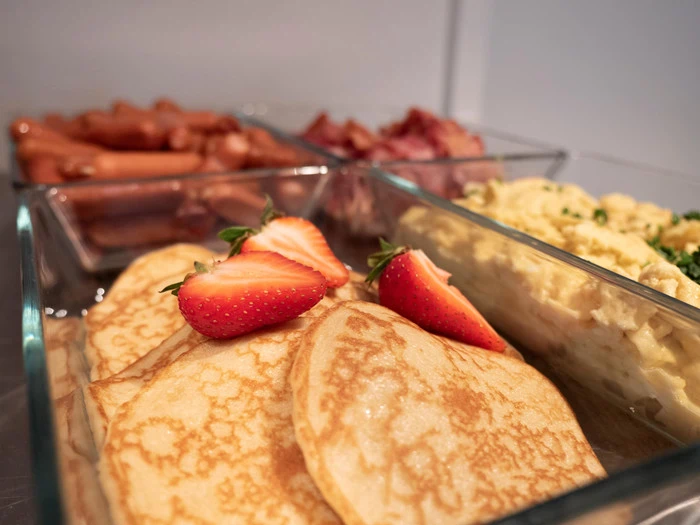 Nærbillede af pandekager toppet med jordbær i skiver, med røræg, bacon og pølser i glasbakker i baggrunden.