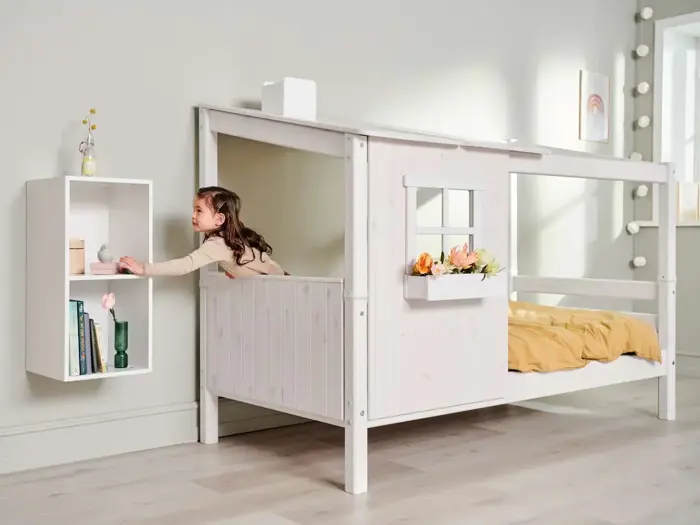 En ung pige i en legehuslignende seng rækker ud efter en væghylde med bøger, en vase og et stykke legetøj. Sengen har et vindue og en blomsterkasse med et gult tæppe indeni.