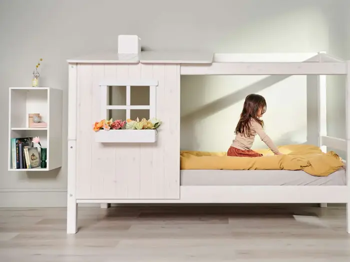En ung pige reder en seng med gule lagner i en hvid træhusformet sengeramme ved siden af en hvid væghylde med bøger og dekorationer.