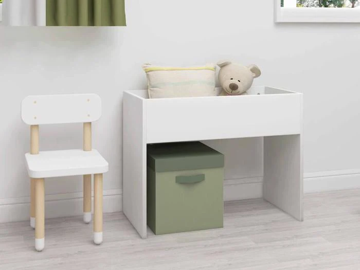 Et lille hvidt skrivebord med en grøn opbevaringsboks under, en pude og en bamse ovenpå, ved siden af en hvid træstol i et lyst, minimalistisk rum.