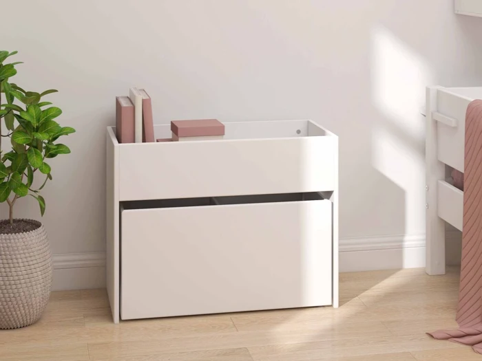 En hvid opbevaringskasse med en nederste skuffe med et par lyserøde bøger står på et trægulv ved siden af en grøn potteplante i et lyst rum.