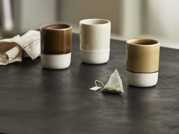 Tre keramiske kopper i jordfarver er placeret på et mørkt bord med en tepose i forgrunden og en foldet klud i baggrunden.