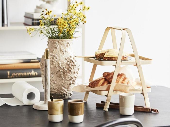 Et træstativ i to etager med kager og et stykke kage står på et bord sammen med tre krus, en tekstureret vase med gule blomster, et stearinlys og bøger i baggrunden.