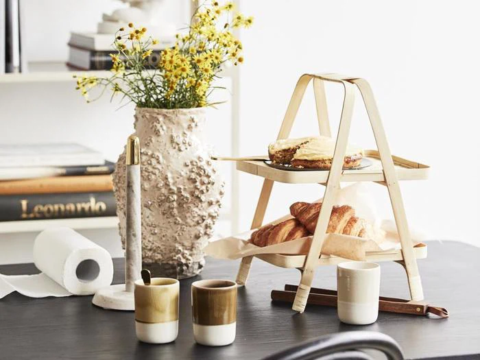 En toetagers træbakke med kager og brød står på et bord med tre keramikkopper, en vase med gule blomster og et par bøger i baggrunden.