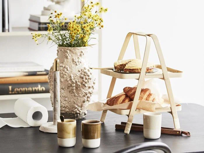 Et træstativ i to etager med kager og et stykke kage står på et bord sammen med tre krus, en tekstureret vase med gule blomster, et stearinlys og bøger i baggrunden.