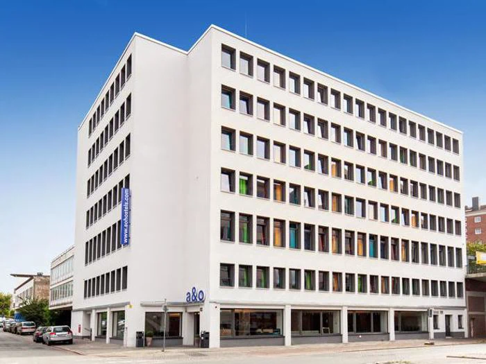 En moderne, hvid bygning i seks etager med mange vinduer og et skilt med teksten a&o ved indgangen mod en klar blå himmel.