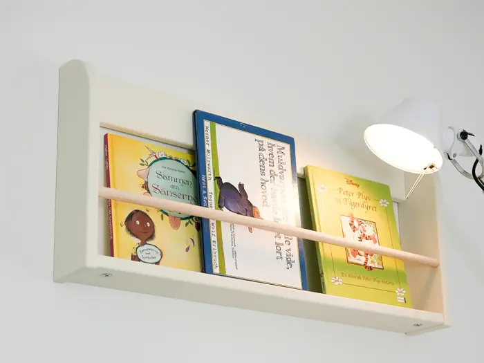 En hvid væghængt bogreol rummer tre børnebøger og oplyses af en hvid lampe, der er monteret på væggen til højre.