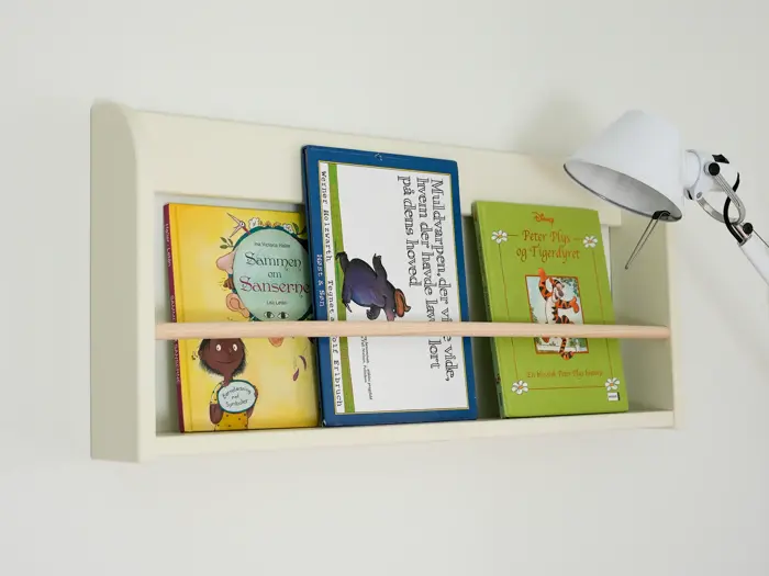 En hvid væghængt hylde rummer fire børnebøger, der holdes fast af en træstang, og en hvid lampe er monteret på hyldens højre side.