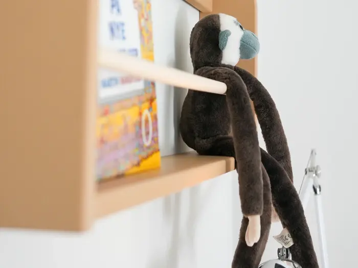 En brun udstoppet abe står på en lys træhylde ved siden af farverige bøger med benene hængende ud over kanten.