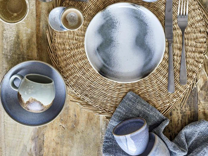 En rustik borddækning med keramiske tallerkener, kopper og skåle i jordfarver på en vævet dækkeserviet, med sølvtøj og en grå serviet på et træbord.
