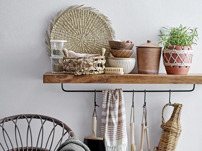 En træhylde med flettede kurve, lerkrukker, træskåle, en potteplante og køkkenredskaber hængende under; en stol er delvist synlig til venstre.