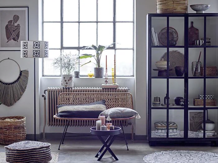 En moderne stue med et stort vindue, kurvemøbler, et sort vitrineskab, pyntepuder, planter og forskellige boligindretningsgenstande i neutrale toner.