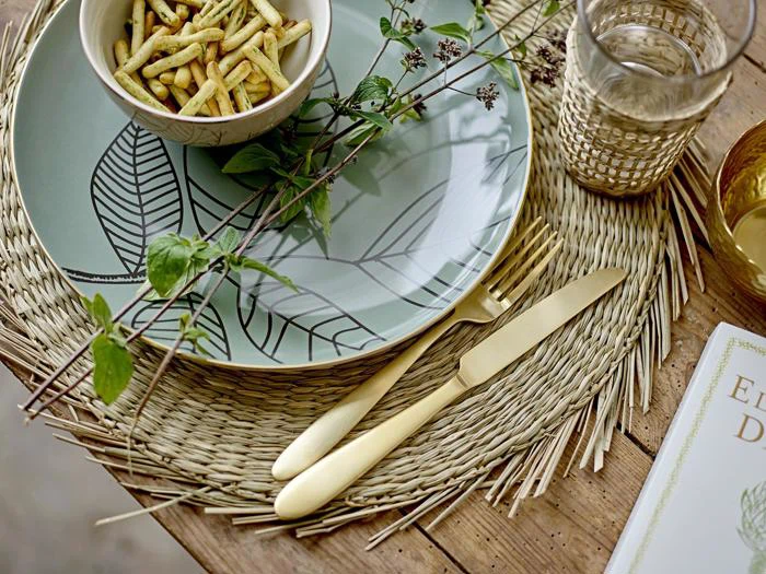 En dekorativ tallerken med bladdesign rummer en lille skål med snacks. Guldbestik, et glas og bladkviste ligger på en vævet dækkeserviet på et rustikt træbord.