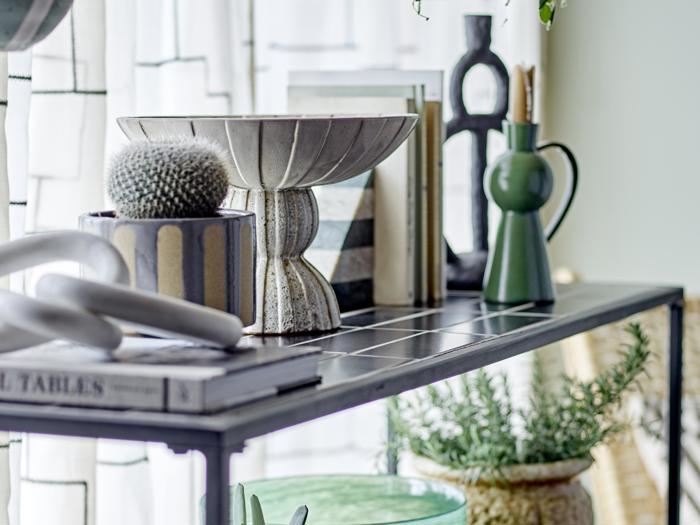 På en moderne hylde står en dekorativ skål, bøger, en kaktus i potte, en grøn vase og en plante, mens sollyset strømmer ind gennem mønstrede gardiner i baggrunden.