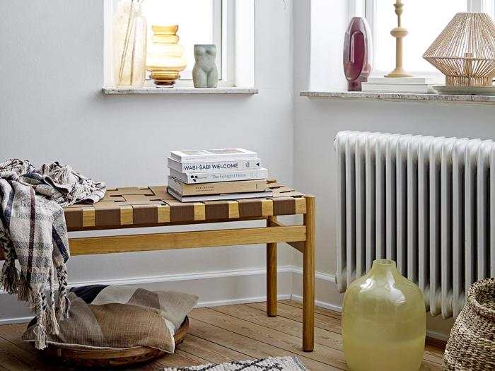 En træbænk med stablede bøger og et tæppe sidder ved et vindue. Dekorative vaser, puder og en lampe fuldender det hyggelige, skandinavisk inspirerede rum.
