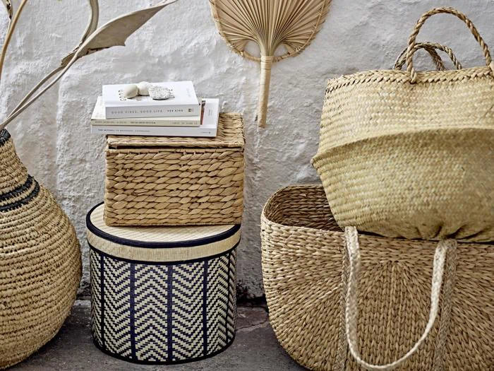 Flettede kurve og kasser i naturlige toner, med stablede bøger og dekorative tørrede palmeblade, arrangeret mod en hvid struktureret væg.