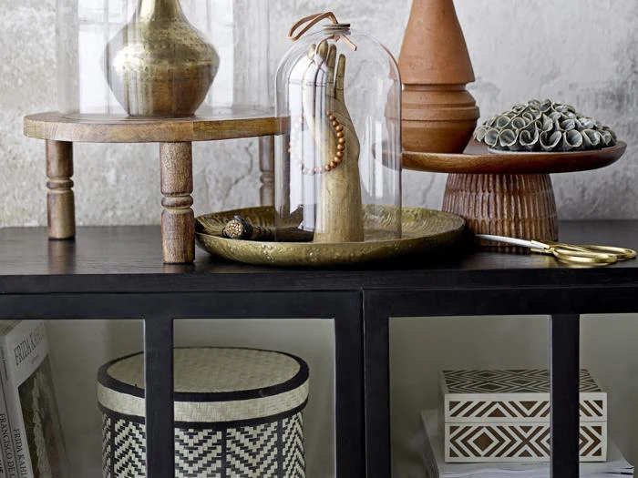 Et sort skab med glasdøre viser dekorative genstande, herunder vaser, bakker, en cloche med en træhånd, perler, lagdelte træstativer og mønstrede æsker.