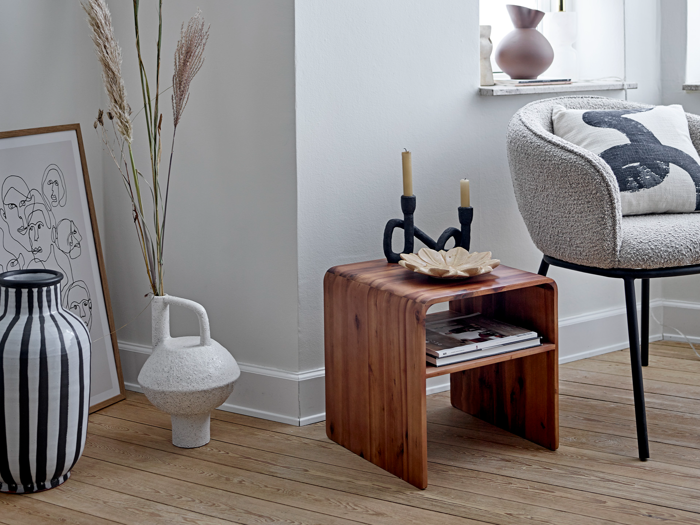 Et hyggeligt, moderne hjørne med et sidebord i træ med sorte lysestager og dekorationer ved siden af en lænestol, vaser, kunstværker og et vindue.