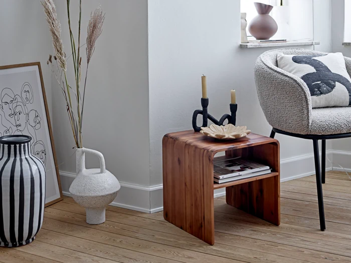 Et hyggeligt, moderne hjørne med et sidebord i træ med sorte lysestager og dekorationer ved siden af en lænestol, vaser, kunstværker og et vindue.