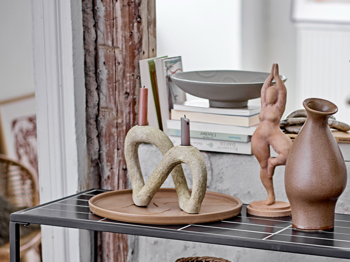 En keramisk bakke rummer to moderne, sløjfeformede lysestager og stearinlys. I nærheden står kunstbøger, en grå skål, en solbrun kande og en lille abstrakt menneskeskulptur på en sort-hvid hylde.