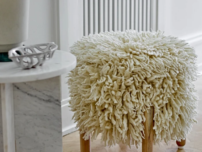 En fluffy, struktureret cremefarvet skammel med træben står ved siden af et rundt marmorbord i et lyst, moderne rum.