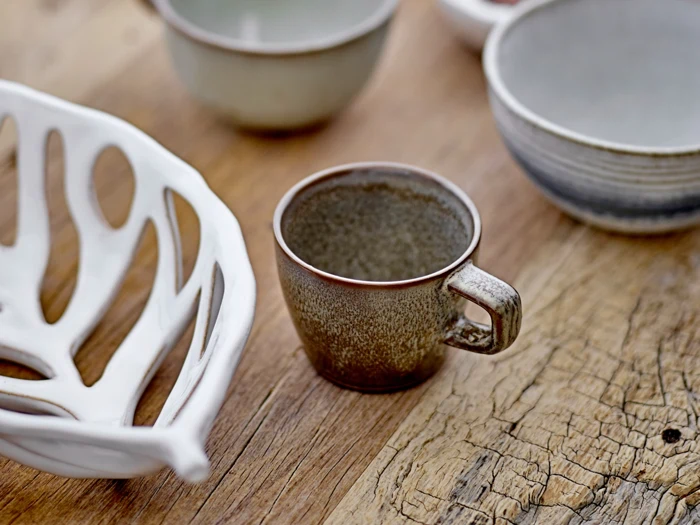 En brun keramikkop står på et træbord, omgivet af et hvidt dekorativt fad og andre keramikskåle.