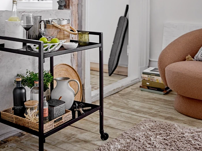 En sort barvogn rummer drinks, glas og en skål med limefrugter i et hyggeligt opholdsrum med en brun stol, et lurvet tæppe og bøger stablet nær en døråbning.