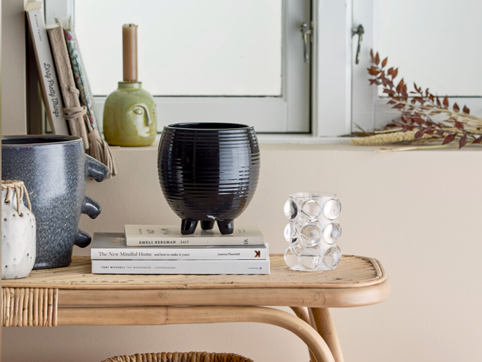 Et rattanbord med dekorative vaser, et glas med boblelignende design, bøger, tørrede blade og et stearinlys foran et hvidt vindue.