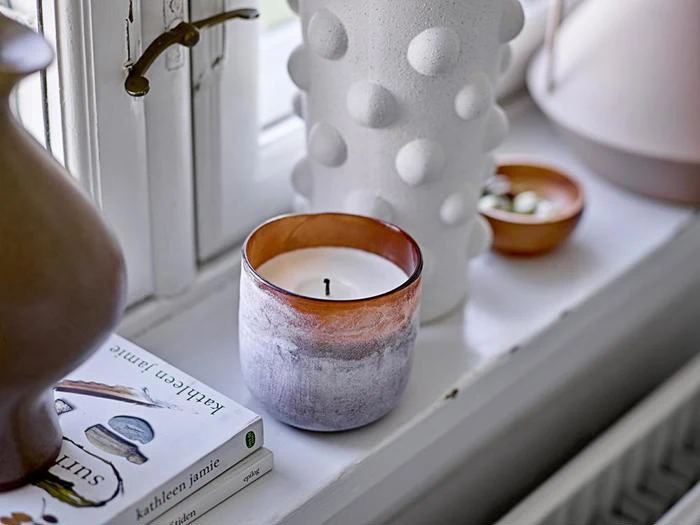 Et stearinlys i en gradientbrun og hvid holder står i vindueskarmen ved siden af en hvid, struktureret vase, en lille skål og to bøger ved et vindue.