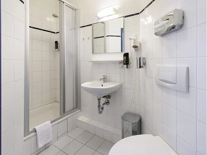 Et lille, hvidt flisebadeværelse med glasbruser, håndvask, spejl, hårtørrer, vægmonterede sæbedispensere, toilet, skraldespand og et håndklæde, der hænger på brusedøren.