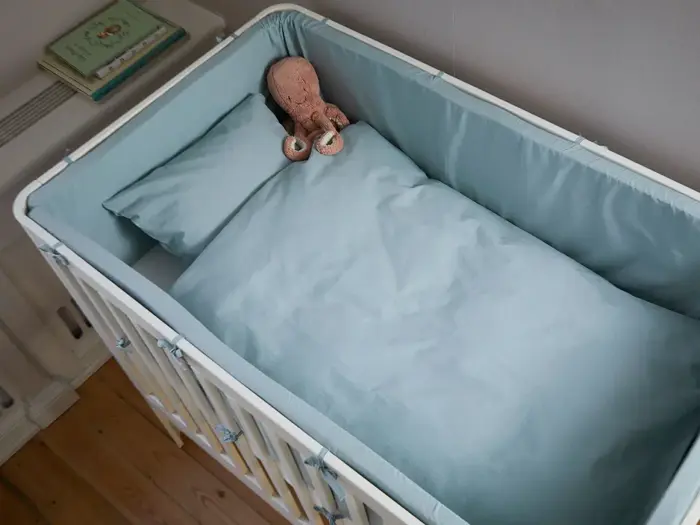 En hvid tremmeseng med lyseblåt sengetøj og en brun, udstoppet blæksprutte på puden, placeret i et svagt oplyst rum med trægulv.