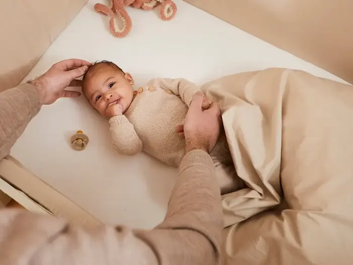 En baby i beige tøj ligger i en krybbe, delvist dækket af et tæppe, mens en voksen forsigtigt rører ved babyens hoved og holder hånden. En sut og et tøjdyr er i nærheden.