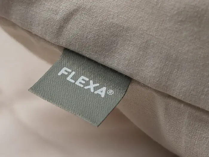 Et nærbillede af et stofmærke mærket FLEXA® syet på beigefarvet sengetøj eller en pude.