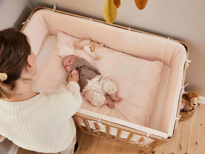 En kvinde står ved siden af en krybbe af træ og holder forsigtigt en baby i hånden, som ligger på en blød, lyserød madras med et tøjdyr i nærheden.