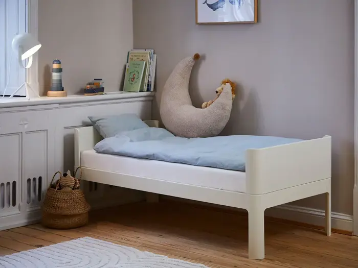 En lille, hvid børneseng med blåt sengetøj, en halvmånepude og en løve som legetøj i et hyggeligt rum med bøger, en lampe og en flettet kurv på et trægulv.