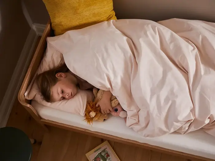 Et lille barn sover i en seng og kæler for et tøjdyr under et lyserødt tæppe med en gul pude og en bog på gulvet i nærheden.